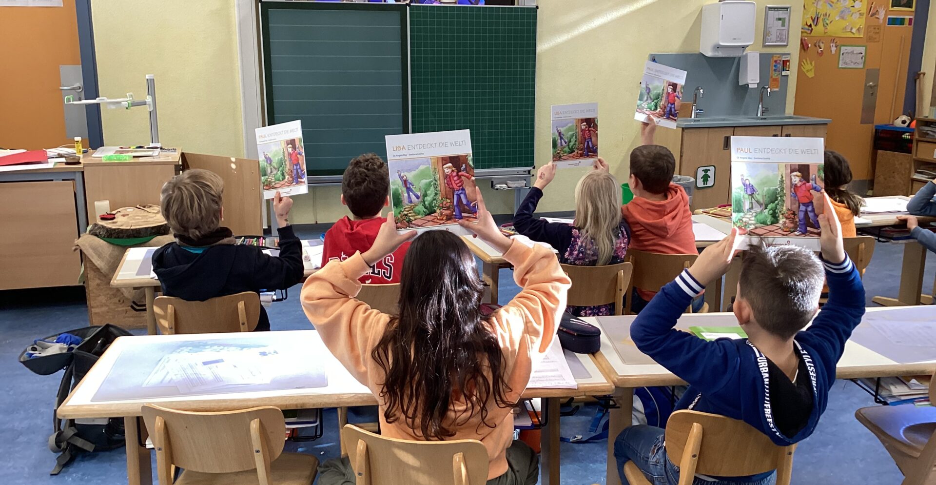 Pupils in a primary school class sit with their backs to the photographer and hold up books.