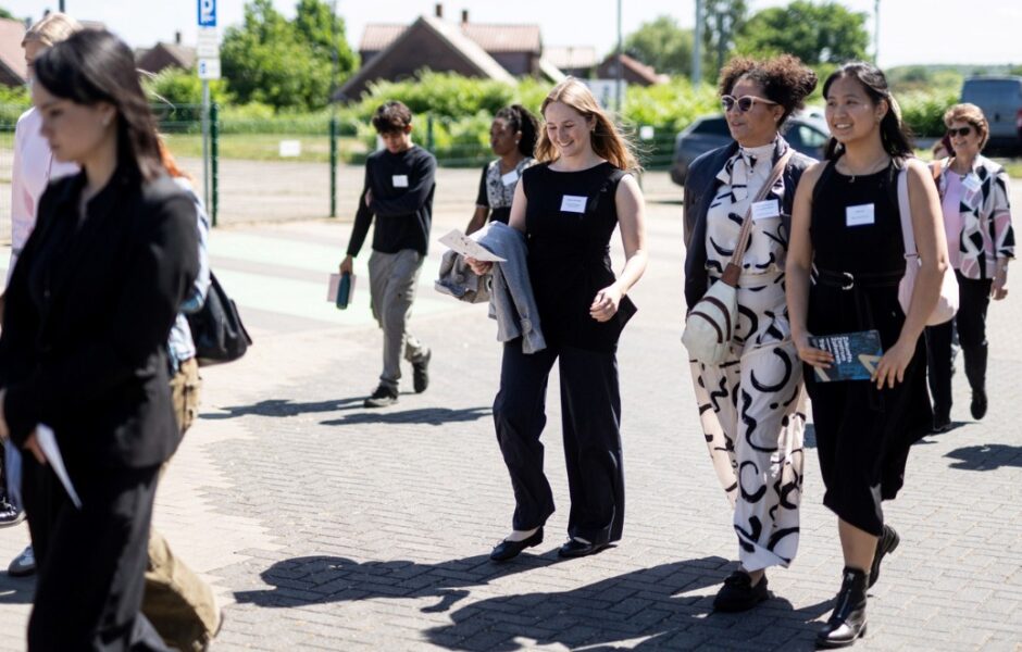 A group of people in formal attire is walking together outdoors on a paved path. The sun is shining, and some individuals are smiling and chatting. In the background, there are residential houses, trees, and a parked van.
