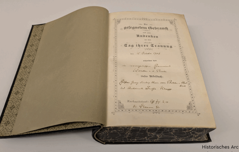 An open Bible lies on a table. The first page reads: ‘In blessed use and in memory of the day of your wedding on 15 October 1906, the priest from Werden a.d. Ruhr gave this Bible book to Gustav Georg Friedrich Maria Krupp von Bohlen und Halbach and Bertha Krupp.’